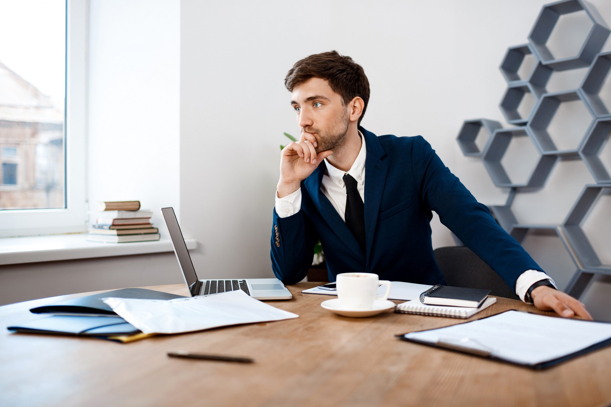 Young successful businessman in suit sitting at workplace, thinking, office background.